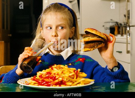 Teenage young schoolgirl eating a huge hamburger and chips junk food she is drinking coke at home in after school London HOMER SYKES Stock Photo