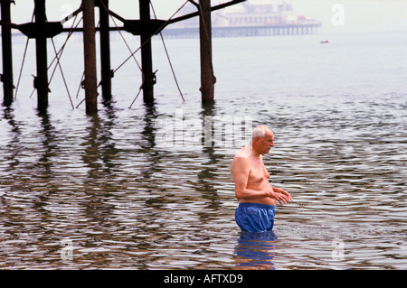 Elderly older man in swimming shorts paddling in the sea with Brighton's two piers in background . Circa 1995 1990s UK HOMER SYKES Stock Photo