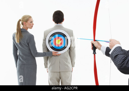 Business woman holding target to man's back while other man (close-up of hands) aims bow and arrow, against white background Stock Photo