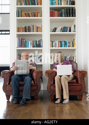 Couple sitting in armchairs, man reading newspaper, woman using laptop Stock Photo