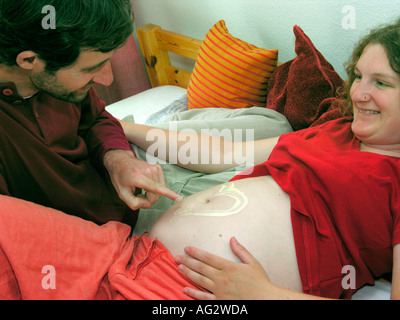laughing pregnant woman lying on a bed letting her partner draw a heart with body oil cream on her belly Stock Photo