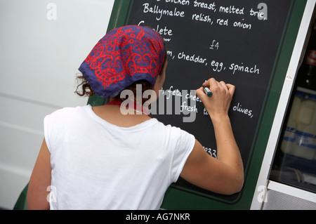 young woman in white t shirt and headscarf writes on blackboard menu with white pen at crepe stall Stock Photo