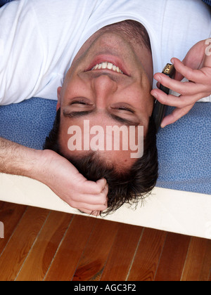 Young man wearing t-shirt stretching on bed talking with mobile phone looking happy looking upwards upside down Stock Photo