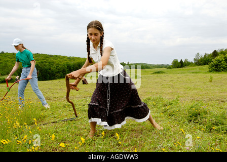 Stock image of a young farm girl in skirt and long braids scything or hand mowing a hay field Stock Photo