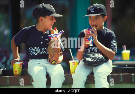 Two young boys wearing baseball uniforms sitting on a wall holding their gloves while eating hot dogs and drinking sodas Stock Photo