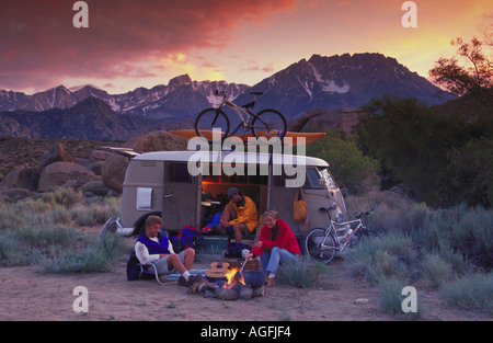 Two men and a woman sitting around a campfire next to a van with mountain bikes and a kayak and mountains in the background