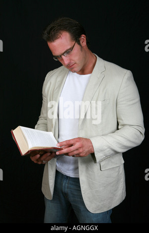 Good looking young man wearing casual sports jacket and glasses reading a large hard backed booked standing up looking serious Stock Photo