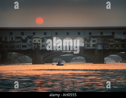 A rowing boat moves  across the river Arno in Florence, as the sun sets over the Ponte Vechio the Old Bridge Stock Photo
