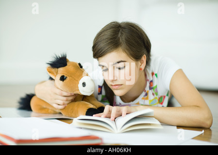 Teen girl lying on floor, holding stuffed toy horse and doing homework Stock Photo