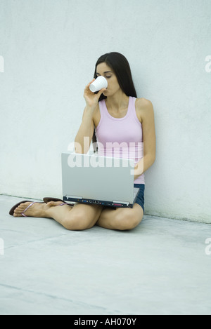 Woman sitting on ground, using laptop and drinking from disposable cup, full length Stock Photo