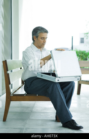 Mature man sitting on bench, opening briefcase, full length Stock Photo