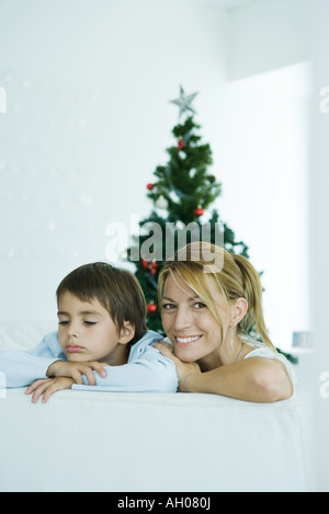 Boy and mother on sofa, Christmas tree in background, woman smiling at camera, boy frowning Stock Photo