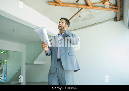 Well-dressed man holding blueprints, using phone, inspecting unfinished home interior Stock Photo