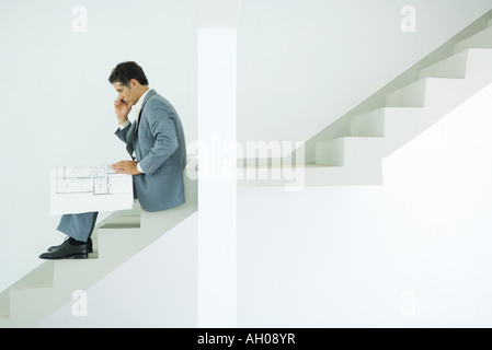 Man in suit sitting on stairs, looking at blueprints, using cell phone Stock Photo