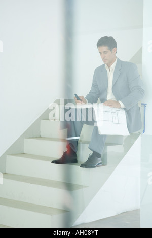 Man in suit sitting on stairs, looking at blueprints, using cell phone, seen through glass Stock Photo