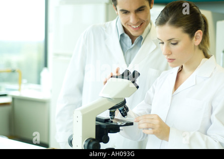 Female lab worker placing slide under microscope, male colleague looking over shoulder, smiling Stock Photo