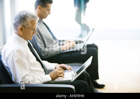 Two businessmen sitting in lobby, both using laptop computers, side view Stock Photo