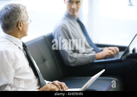 Two businessmen sitting on sofa, looking at each other, both using laptops computers Stock Photo