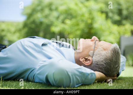 Mature businessman lying on ground outdoors, hands behind head, eyes closed, close-up Stock Photo