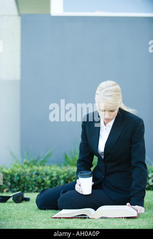 Young businesswoman sitting on the ground outdoors, reading book, holding hot beverage Stock Photo