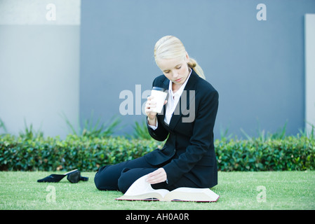 Young businesswoman sitting on the ground outdoors, reading book, holding hot beverage Stock Photo