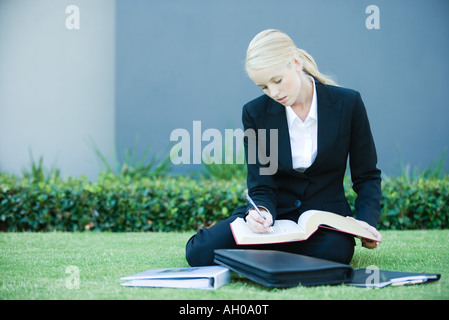 Young businesswoman sitting on the ground outdoors, reading book, holding pen Stock Photo