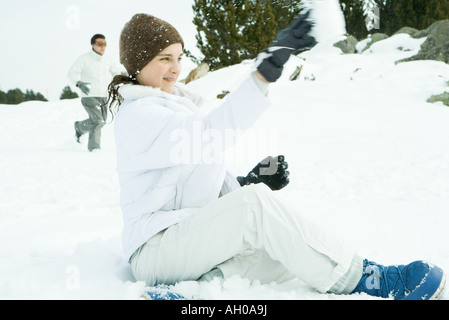 Teenage girl sitting on ground in snow, throwing snowball, side view Stock Photo