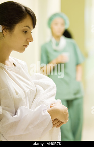 Young woman holding towel in hospital corridor, looking down, medical worker in background Stock Photo