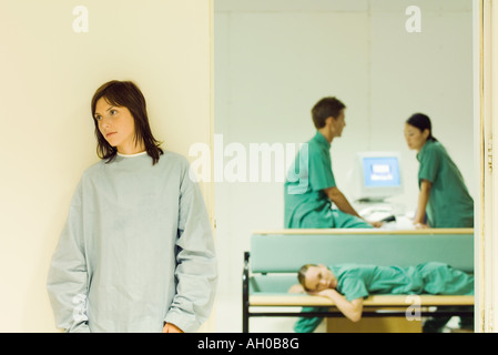 Female patient standing in hospital corridor, medical staff in office in background Stock Photo