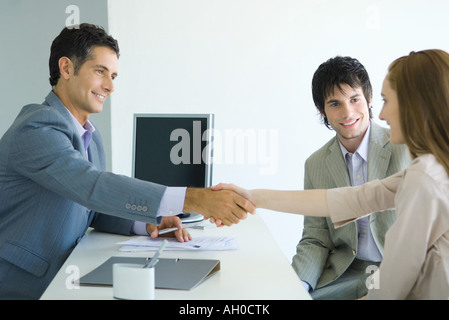 Businessman shaking hands with young woman across desk, young woman's male companion smiling Stock Photo