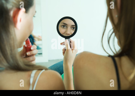 Teenage girl sitting next to friend, applying lipstick, looking at self in hand mirror, rear view Stock Photo