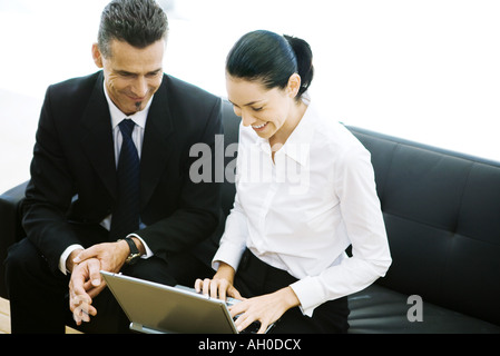 Male and female business associates sitting in lobby, using laptop, smiling Stock Photo