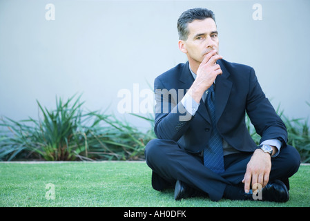 Businessman sitting cross-legged on grass, fingers over lips Stock Photo