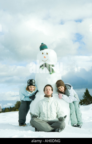 Young man meditating in front of snowman, two sisters crouching behind, smiling at camera Stock Photo