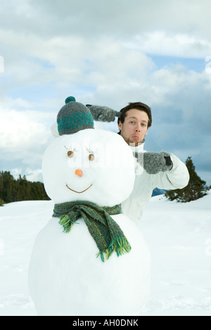 Young man standing behind snowman, pointing, making faces at camera Stock Photo