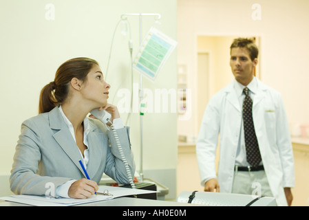 Woman using phone in doctor's office, looking over shoulder at male doctor Stock Photo