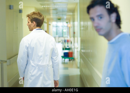 Male doctor walking in hospital corridor, rear view, man looking at camera in foreground Stock Photo