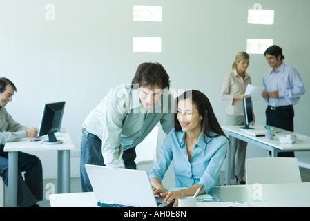 Businessman and businesswoman in office, using laptop computer, associates in background Stock Photo