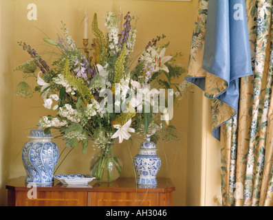 Close-up of white lilies and blue delphiniums in vase on chest with blue+white vases beside blue floral swagged curtains Stock Photo