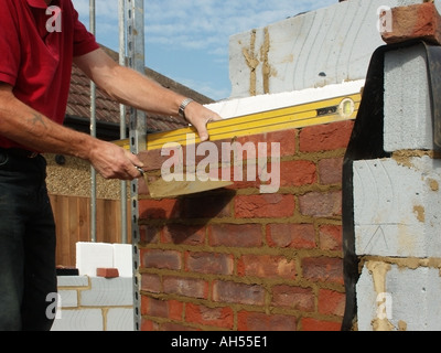 Building construction work in progress close up bricklayer using trowel & spirit level working on facing brick skin of cavity wall house building UK Stock Photo
