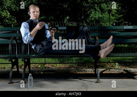 Businessman reclining on a park bench and eating food with chopsticks Stock Photo