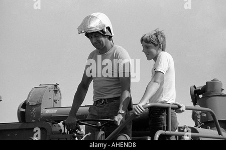 Man driving old model combine harvester wearing Airstream dust protection helmet and carrying a young boy passenger Stock Photo