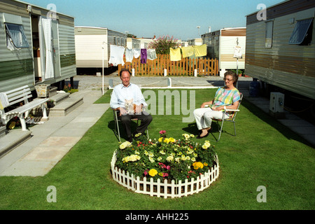 Couple sitting in the sun on grass lawn next to small flowerbed outside their static caravan Trecco Bay Porthcawl South Wales UK Stock Photo