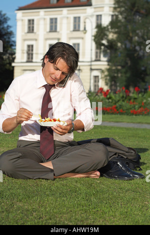 Businessman on square sitting in meadow on the mobile whilst eating chips laughing. Stock Photo