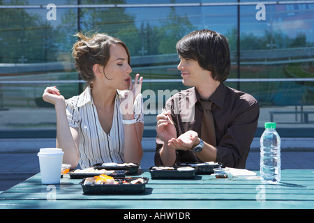 Businessman and woman on table in front of office outside with sushi lunchbox and sticks she is eating he is watching her. Stock Photo