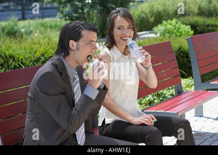 businessman and woman sitting on a bench outdoors she's about to drink water he is having a sandwich Stock Photo