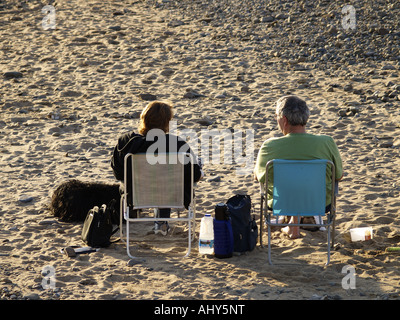 Middle aged couple sitting on deck chairs on a sandy beach early evening, looking out at the sea Stock Photo
