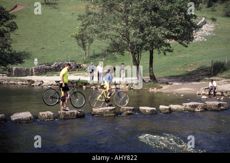 Cyclists carrying bikes crossing River Dove stepping stones in  picturesque Dovedale National Nature Reserve Peak District Derbyshire Staffordshire UK Stock Photo