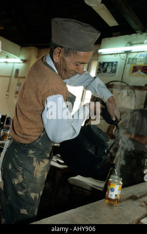 Elderly Hindu man wearing a cap and vest pouring hot tea from a large steaming black kettle into a glass jar in a workshop. Stock Photo