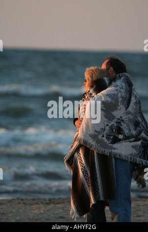 Couple standing on beach wrapped in blankets looking at the sun Stock Photo